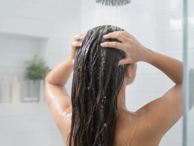 A person rinsing shampoo out of long dark hair under a shower head in a bright tiled shower.